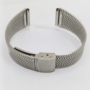 Puede incluir: Una correa de reloj plateada, de estilo malla, con cierre desplegable. La correa está hecha de pequeños eslabones metálicos interconectados, creando una superficie flexible y texturizada. El cierre es rectangular con cierre de bisagra.