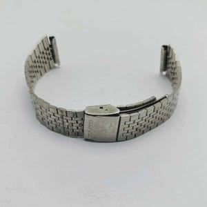 May include: A silver-toned watch band with a woven metal design. The band features a clasp with the brand name "CASIO" engraved on it. The band is curved and ready to be attached to a watch.