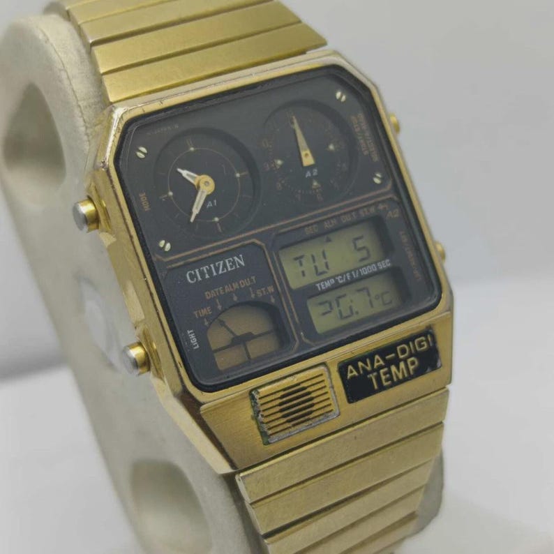 May include: A gold-coloured Citizen Ana-Digi Temp watch. The watch has a black face with both analogue and digital displays, showing the date, time, and temperature. The watch strap is also gold-coloured.