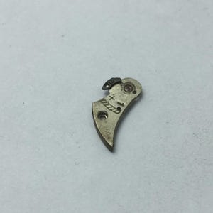 May include: A small, curved, metallic watch part with a plus and minus symbol etched on its surface. The part has a small hole and a curved shape, with the word "ROW" etched on the bottom. A small, black piece is attached to the top.