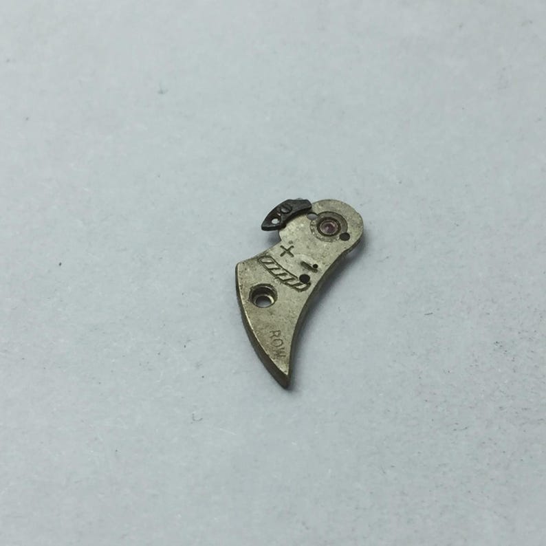 May include: A small, curved, metallic watch part with a plus and minus symbol etched on its surface. The part has a small hole and a curved shape, with the word "ROW" etched on the bottom. A small, black piece is attached to the top.