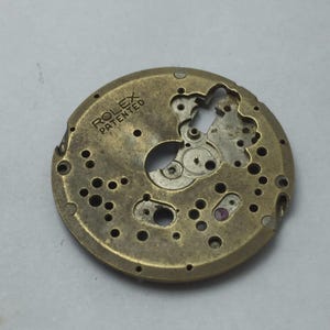 May include: A vintage Rolex watch movement component. The circular, brass-colored part features intricate cutouts, holes, and gears. The text "ROLEX PATENTED" is stamped on the surface. A small, red jewel is visible.