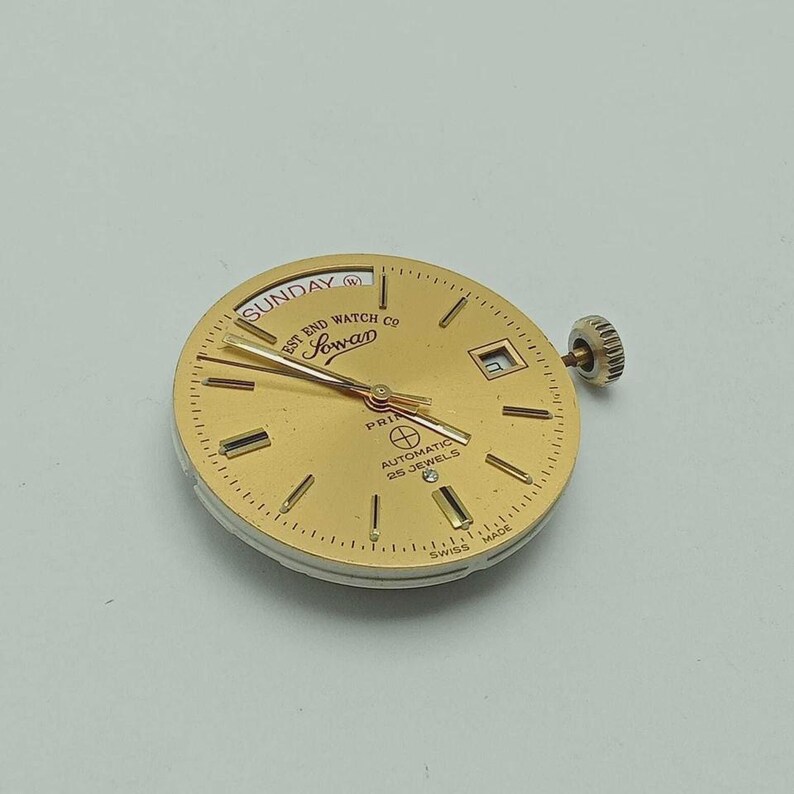 May include: A gold-toned watch face with the text "Sunday" and "West End Watch Co." The watch face has gold hands and markers, a date window, and the text "Sowan". The watch is marked "Automatic 25 Jewels" and "Swiss Made".