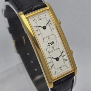 May include: A rectangular gold-toned wristwatch with two white watch faces and black hands. The top face displays Arabic numerals, while the bottom face has Roman numerals. The watch has the text "ANA" and a black leather strap.