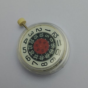 Puede incluir: Un componente de reloj con una esfera blanca con números negros y una esfera interior roja con números. Una pequeña perilla de color crema está unida a la parte superior. El componente está encerrado en una carcasa transparente y circular.
