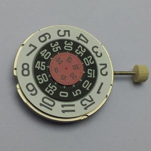 May include: A watch dial with a white outer ring featuring black numerals from 1 to 12. The dial has a black inner ring with numbers from 5 to 55 in increments of 5. A smaller red ring displays numbers from 0 to 50. A small beige crown is attached.