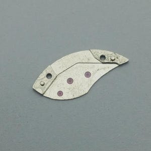 K&ouml;nnte beinhalten: Ein silberfarbenes Uhrenteil mit einer gebogenen, asymmetrischen Form. Es hat mehrere kleine L&ouml;cher und drei rosa kreisf&ouml;rmige Akzente. Das Teil scheint aus Metall zu sein und hat eine leicht strukturierte Oberfl&auml;che.