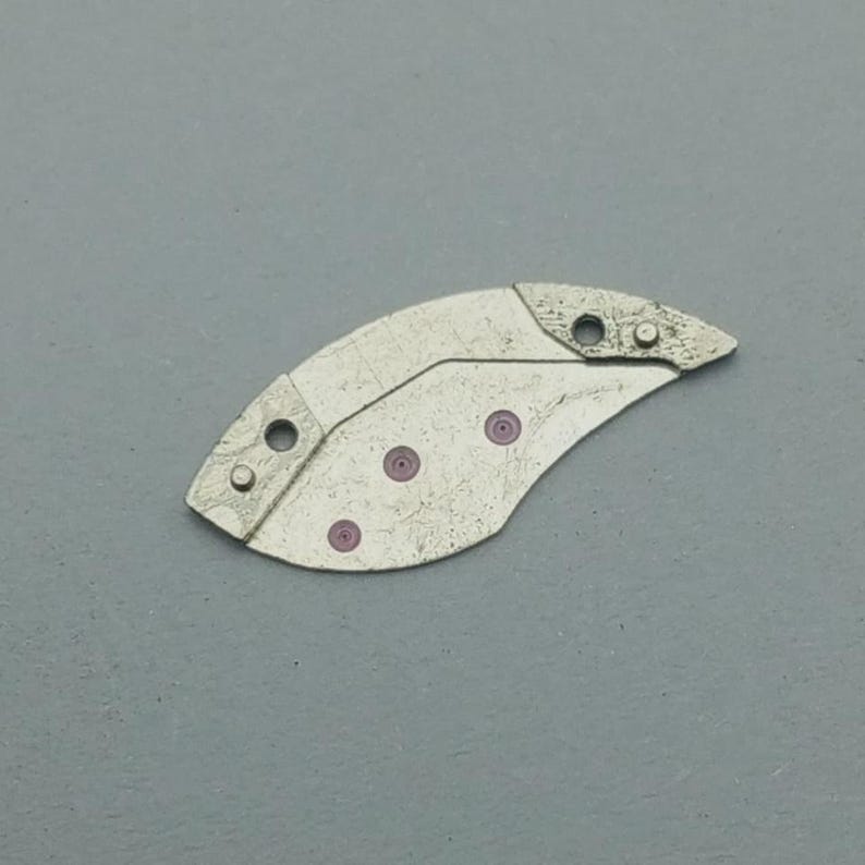 K&ouml;nnte beinhalten: Ein silberfarbenes Uhrenteil mit einer gebogenen, asymmetrischen Form. Es hat mehrere kleine L&ouml;cher und drei rosa kreisf&ouml;rmige Akzente. Das Teil scheint aus Metall zu sein und hat eine leicht strukturierte Oberfl&auml;che.