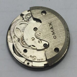 May include: A detailed view of a vintage OMAX watch movement. The silver mechanism displays intricate gears, a balance wheel, and the text "17 SEVENTEEN JEWELS" and "SWISS". The OMAX logo is visible on the upper right.