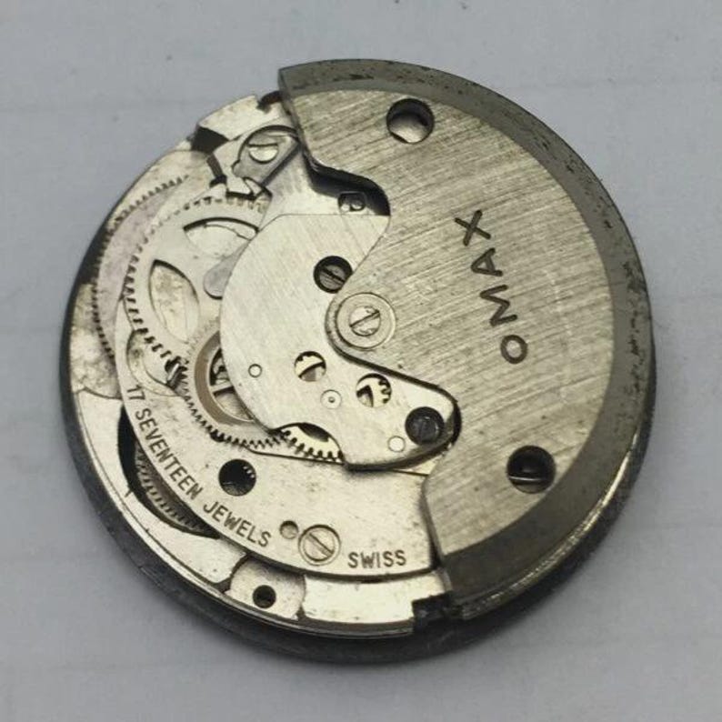 May include: A detailed view of a vintage OMAX watch movement. The silver mechanism displays intricate gears, a balance wheel, and the text "17 SEVENTEEN JEWELS" and "SWISS". The OMAX logo is visible on the upper right.