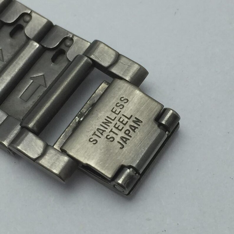 Puede incluir: Primer plano de un cierre de correa de reloj de acero inoxidable. El cierre es plateado y tiene las palabras "STAINLESS STEEL JAPAN" impresas. La correa est&aacute; hecha de m&uacute;ltiples eslabones met&aacute;licos.