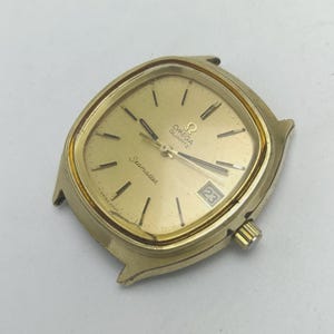 May include: A gold-coloured Omega Seamaster Quartz watch face with a square design. The watch face has a gold dial with black hour markers and hands. The date is displayed at the 3 o'clock position. The watch is marked "Swiss Made".