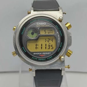 Op de afbeelding: Een zilveren Casio digitale horloge met een zwarte band. De wijzerplaat toont de tijd, datum en andere functies. Het horloge heeft goudkleurige knoppen en de tekst "CASIO" en "SHOCK RESIST" zijn zichtbaar.