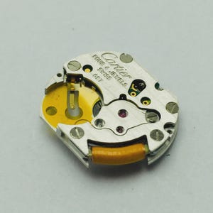 May include: A close-up of a watch movement with the Cartier logo and the text "FOUR & JEWELS SWISS C57". The movement has a silver metal finish with gold and yellow components, and a small red jewel.