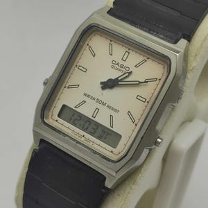 May include: A vintage Casio Quartz watch with a rectangular silver-tone case and a black band. The watch face is off-white with black hands and markers. The digital display shows the time as 12:03. The watch is water resistant.