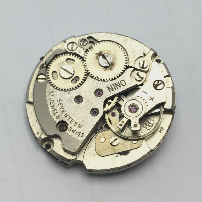 May include: A detailed view of a vintage watch movement, showcasing intricate gears, screws, and jewels. The circular mechanism features the text "NINO" and "SEVENTEEN JEWELS SWISS". The metal parts are silver and gold in colour.