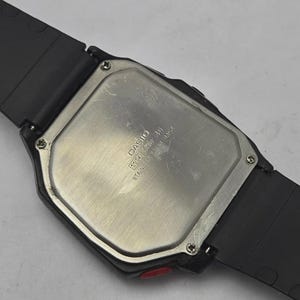 Puede incluir: Primer plano de un reloj digital Casio negro. El reloj tiene una parte trasera de acero inoxidable con el nombre de la marca y el n&uacute;mero de modelo. La correa del reloj es negra y el reloj tiene un bot&oacute;n rojo en el lateral.