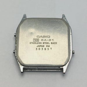 May include: A silver-toned Casio watch back with the text "CASIO", "189 SA-61", "STAINLESS STEEL BACK", "JAPAN NM", and "303957". The watch back has a rectangular shape with rounded corners and a stainless steel finish.