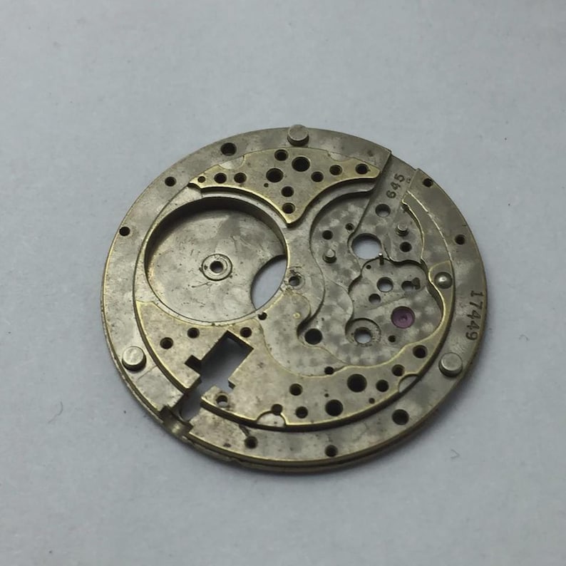 May include: A vintage watch movement with intricate gears and a circular design. The metal components are a mix of silver and gold tones, with small holes and a few visible jewels. The numbers "645" and "17449" are etched on the surface.