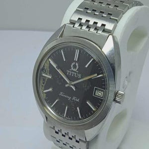 Op de afbeelding: Een zilverkleurig Titus Tuning Fork horloge met een donkergrijze wijzerplaat en een datumweergave op 3 uur. Het horloge heeft een zilveren band en wordt gepresenteerd op een witte standaard. De wijzerplaat toont de merknaam en de woorden "Tuning Fork".