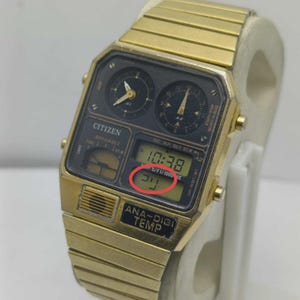 May include: A gold-coloured Citizen Ana-Digi Temp watch. The black watch face features analog dials and a digital display showing the time and temperature. The watch strap is also gold-coloured.
