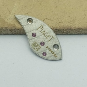 K&ouml;nnte beinhalten: Ein silberfarbenes Uhrenbauteil mit dem Markennamen "Piaget" aufgedruckt. Das Bauteil hat eine gebogene Form mit mehreren kleinen L&ouml;chern und kreisf&ouml;rmigen rosa Akzenten. Der Text "9P2" und "9104524" ist ebenfalls sichtbar.
