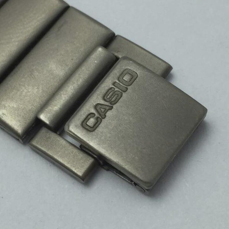 Puede incluir: Primer plano de una secci&oacute;n de una correa de reloj. La correa es de metal gris mate. El cierre es cuadrado con el nombre de la marca "CASIO" en relieve. Los eslabones de la correa son rectangulares y est&aacute;n conectados.