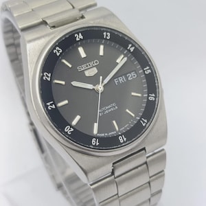 May include: A silver-toned Seiko 5 automatic wristwatch with a gray face and a black bezel. The watch displays the day and date, with the day reading "FRI 25". The watch has a silver band and the text "AUTOMATIC 21 JEWELS" is visible.