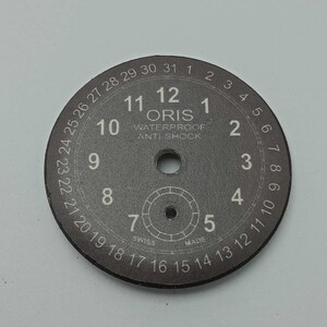 May include: A round, dark gray watch dial featuring white Arabic numerals from 1 to 12, a date track numbered 1 to 31 along the outer edge, and the text ORIS WATERPROOF ANTI-SHOCK printed in the center with SWISS MADE at the bottom.
