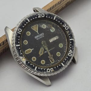 Peut inclure: Une montre Seiko Quartz vintage avec un cadran noir et une lunette argentée. Le cadran affiche le jour "TUE 25". La lunette a des marquages blancs et la montre a des cornes argentées.