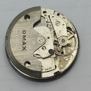 May include: A detailed view of an OMAX watch movement, showcasing its internal components. The silver-toned mechanism features gears, screws, and a ruby bearing. The word "OMAX" is visible, with "SWISS" at the base.