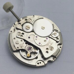 May include: Close-up of a silver watch movement with intricate gears, screws, and a winding stem. The watch mechanism has a circular design with the word "SWISS" visible. The overall theme is horology and watchmaking.