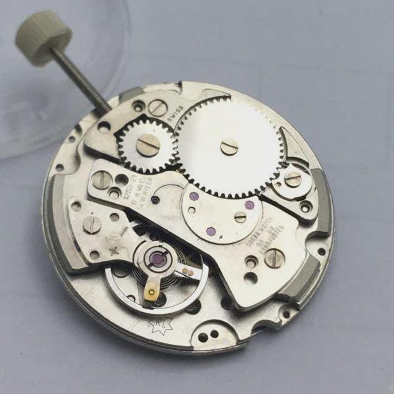 May include: Close-up of a silver watch movement with intricate gears, screws, and a winding stem. The watch mechanism has a circular design with the word "SWISS" visible. The overall theme is horology and watchmaking.