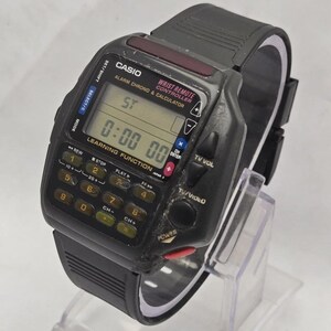 Puede incluir: Un reloj Casio Wrist Remote Controller vintage. El reloj negro presenta un teclado de calculadora, una pantalla digital que muestra "0:00 00" y botones de funci&oacute;n. La correa del reloj es de goma negra.