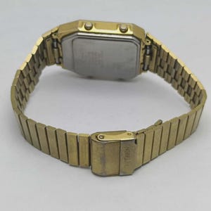 May include: A gold-toned Citizen digital watch with a rectangular face and a matching metal band. The watch has a clasp closure and a silver-coloured back. The band is open, showing the inside of the watch.
