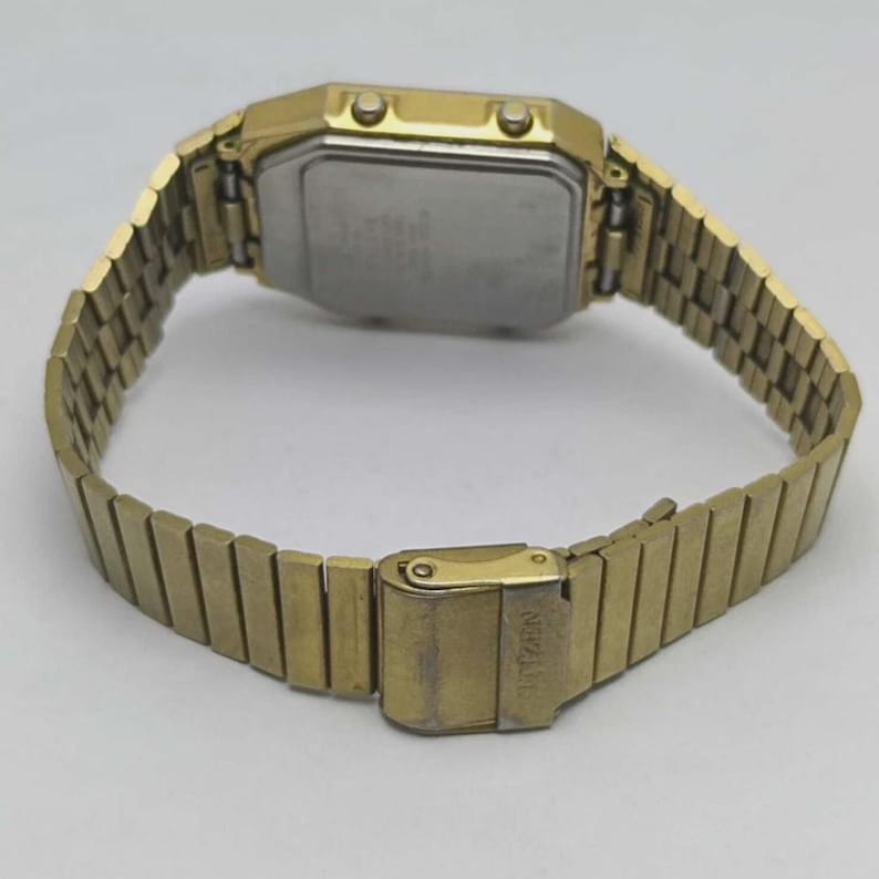 May include: A gold-toned Citizen digital watch with a rectangular face and a matching metal band. The watch has a clasp closure and a silver-coloured back. The band is open, showing the inside of the watch.