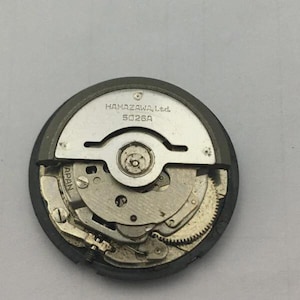 Może przedstawiać: Zbliżenie mechanizmu zegarka z widocznymi kołami zębatymi i elementami. Metalowy mechanizm ma napis "HAMAZAWA, Ltd. 5026A" wydrukowany na srebrnej płytce. Mechanizm zegarka jest okrągły i ma ciemnoszarą obw&oacute;dkę.