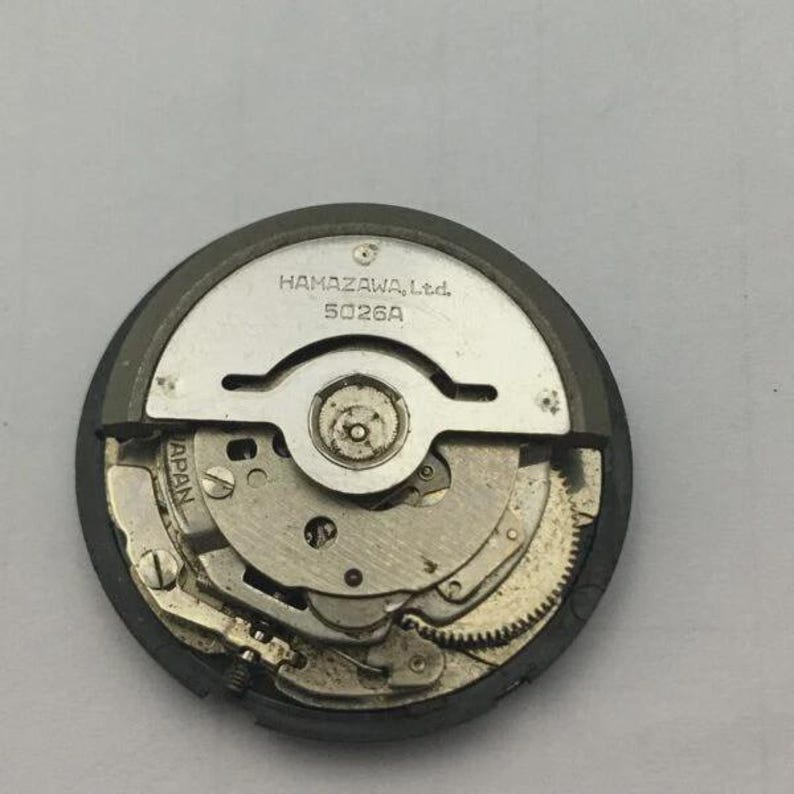 Może przedstawiać: Zbliżenie mechanizmu zegarka z widocznymi kołami zębatymi i elementami. Metalowy mechanizm ma napis "HAMAZAWA, Ltd. 5026A" wydrukowany na srebrnej płytce. Mechanizm zegarka jest okrągły i ma ciemnoszarą obw&oacute;dkę.
