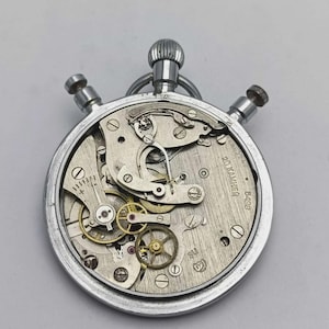Könnte beinhalten: Eine silberne Vintage-Stoppuhr mit freiliegenden Innenrädern und Mechanismen. Das Zifferblatt zeigt komplizierte Details des Innenlebens, einschließlich kleiner gold- und silberfarbener Komponenten. Die Oberseite hat eine Aufzugskrone und zwei Seitentasten.