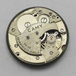 May include: Close-up of a vintage CAMY watch movement. The mechanism features numerous gears, jewels, and the text "CAMY", "SWISS", and "SEVENTEEN 17 JEWELS". The metal components are silver and gold-toned, with a circular shape.