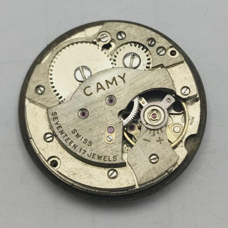 May include: Close-up of a vintage CAMY watch movement. The mechanism features numerous gears, jewels, and the text "CAMY", "SWISS", and "SEVENTEEN 17 JEWELS". The metal components are silver and gold-toned, with a circular shape.