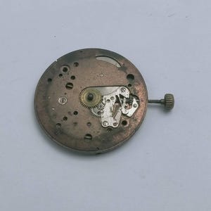 May include: A close-up of a vintage watch movement. The circular, bronze-colored mechanism features intricate gears, levers, and screws. A small, textured knob extends from the side. The watch movement is set against a plain, light background.