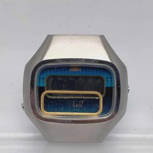 Könnte beinhalten: Ein silberfarbenes Digitaluhrengehäuse mit blauem Zifferblatt und einem goldfarbenen, rechteckigen Rahmen. Auf dem Display ist der Text "INT" und "ARTZ" in Weiß aufgedruckt. Das Uhrengehäuse hat eine glatte, metallische Oberfläche und Form.
