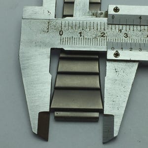 Puede incluir: Primer plano de un eslab&oacute;n de correa de reloj de metal que se mide con un calibrador de metal. El eslab&oacute;n es de color plata apagado y tiene un dise&ntilde;o escalonado. El calibrador muestra medidas.