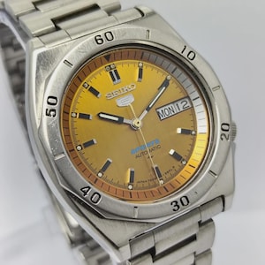 Puede incluir: Un reloj automático Seiko 5 Sports de acero inoxidable con esfera dorada. La esfera muestra el día y la fecha, con una pista de minutos alrededor del borde exterior. La correa del reloj también es de acero inoxidable.