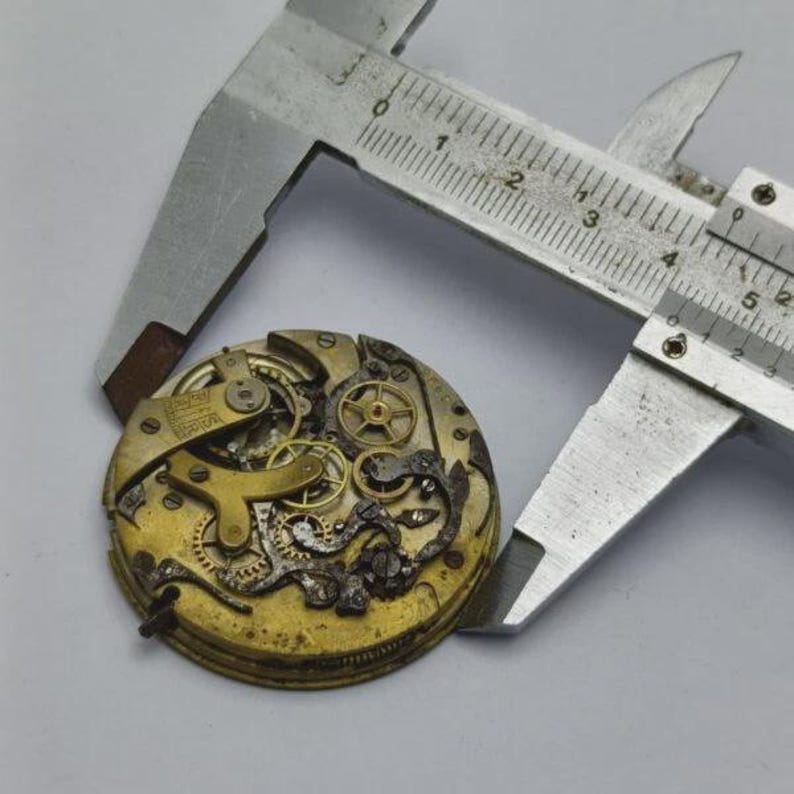 May include: Close-up of a vintage watch movement with intricate gears and components, held by a silver caliper. The watch mechanism has a gold and brass color, showing signs of age and wear. The caliper is used to measure the watch's diameter.
