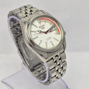 Pu&ograve; includere: Un orologio da polso automatico Seiko 5 color argento con quadrante bianco e cinturino in acciaio inossidabile. Il quadrante mostra il giorno e la data, con dettagli rossi e bianchi. L'orologio &egrave; su un espositore trasparente.