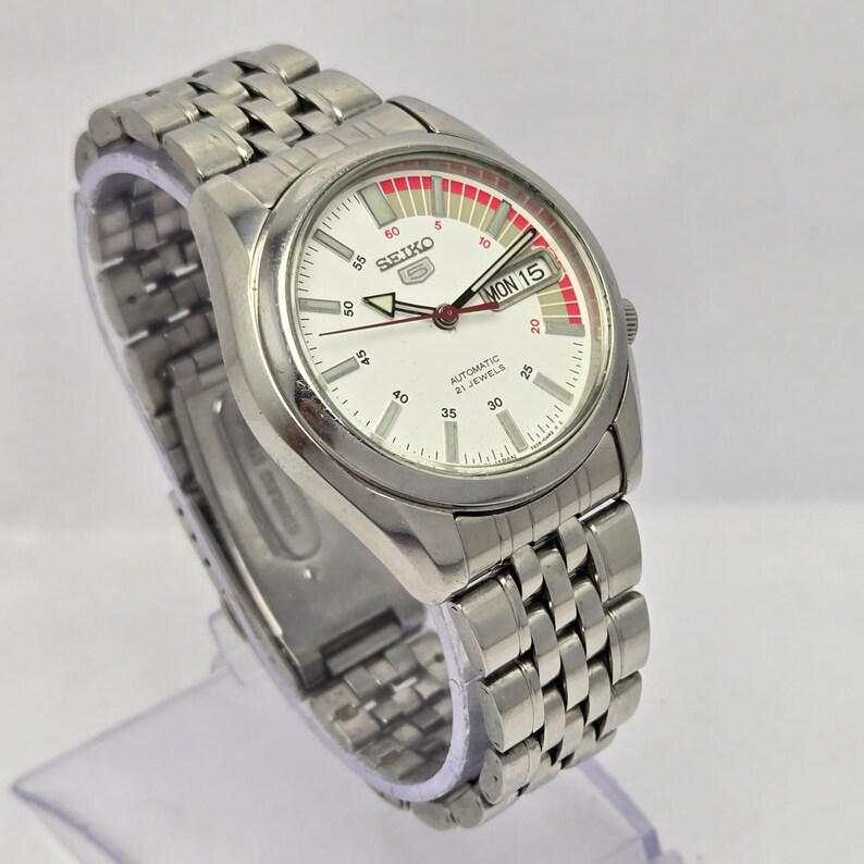 Pu&ograve; includere: Un orologio da polso automatico Seiko 5 color argento con quadrante bianco e cinturino in acciaio inossidabile. Il quadrante mostra il giorno e la data, con dettagli rossi e bianchi. L'orologio &egrave; su un espositore trasparente.