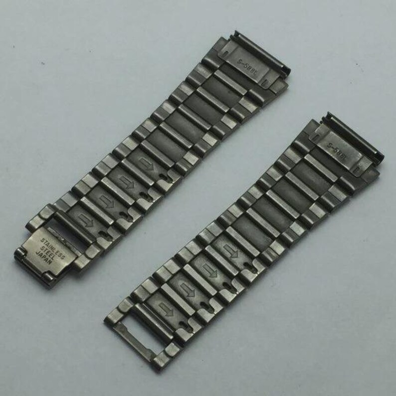 Puede incluir: Dos correas de reloj de acero inoxidable con un acabado de metal cepillado. Cada correa tiene una serie de eslabones rectangulares con flechas direccionales y el texto "STAINLESS STEEL JAPAN". Las correas est&aacute;n dise&ntilde;adas para un reloj.