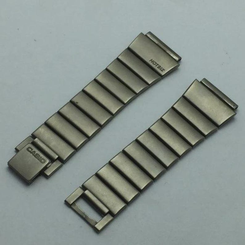 Puede incluir: Dos correas de reloj plateadas. Una correa tiene un cierre con la palabra "CASIO" grabada. La otra correa tiene la palabra "HOTBIZ" grabada. Ambas correas est&aacute;n hechas de eslabones met&aacute;licos rectangulares.